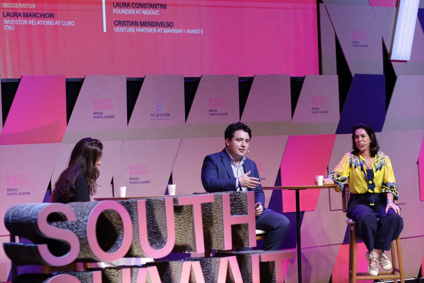 Cristian Mendivelso participa en un panel de South Summit Brazil, sentado al centro del escenario rosa junto a otras dos panelistas.