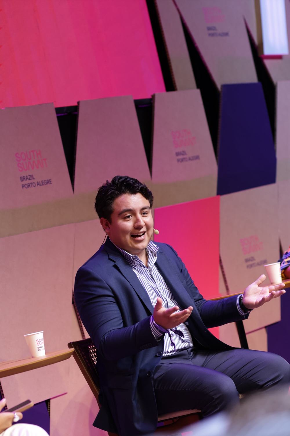 Cristian Mendivelso habla con las manos abiertas durante un panel en South Summit Brazil, frente al fondo rosa del escenario.